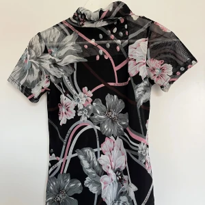Y2K Blommig mesh polotopp  - Säljer en svart mesh polotopp från Pineapple med stora blommor i grått, rosa och vitt. Toppen har korta ärmar och en liten polokrage. Materialet är genomskinligt och stretchigt, perfekt för att sticka ut. Skulle säga att storleken är S/M, den är stretchig. 