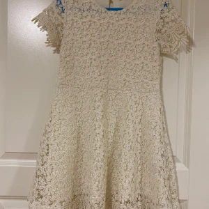 Vit spetsklänning med kort ärm - Söt vit klänning i spets med korta ärmar och rund halsringning. Klänningen har ett vackert spetsmönster över hela plagget och en lätt utställd kjol. Perfekt för sommarens tillfällen eller student.