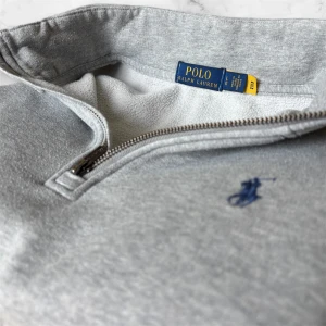 Grå halvzip tröja från Polo Ralph Lauren - Snygg grå tröja från Polo Ralph Lauren med halv dragkedja och broderad marinblå logga på bröstet. Tröjan har lång ärm, hög krage och är tillverkad i mjuk bomullsmix. Perfekt för en avslappnad och stilren look.