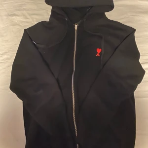 Svart hoodiejacka med dragkedja från Ami Paris - Svart hoodiejacka från Ami Paris med dragkedja framtill och röd broderad logga på bröstet. Jackan har huva och en enkel, stilren design som passar perfekt till vardags. Klassisk passform och mjukt material.