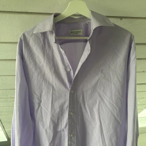 Ljuslila skjorta från Yves Saint Laurent - Säljer en stilren ljuslila skjorta från Yves Saint Laurent med klassisk krage och diskret YSL-broderi på bröstet. Skjortan har långa ärmar och knäppning framtill. Perfekt för dig som gillar en clean och exklusiv look. Den är i mycket bra skick, använd ca 4 gånger. Enda defekten är att kragen längst ut på armen gått upp lite (syns på sista bilden) storlek M Hör gärna av er vid frågor och funderingar!