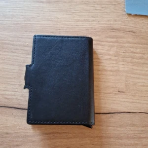 Svart plånbok i skinn - Säljer en stilren svart plånbok i skinn med enkel design. Plånboken har en knappstängning på sidan och plats för kort. Perfekt för dig som vill ha en smidig och klassisk accessoar i fickan.