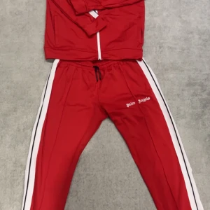 Palm angles red tracksuit Storlek XL - Röda träningsbyxor från Palm Angels med vita revärer längs sidorna och svart dragsko i midjan. Byxorna har logotypen broderad på vänster sida och är tillverkade i polyester och elastan för en skön och sportig känsla.
