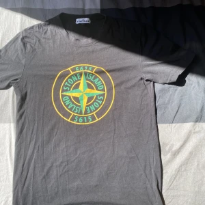 Grå t-shirt från Stone Island med tryck - Säljer en grå t-shirt från Stone Island med stort gult och grönt logotryck på bröstet. Klassisk passform och rund halsringning. Perfekt för dig som gillar streetwear och vill ha en snygg och enkel t-shirt med tydlig logga.