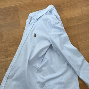 Ljusblå skjorta från U.S. Polo Assn. - Klassisk vit skjorta med broderad logga på bröstet från U.S. Polo Assn. Skjortan har knappar framtill och lång ärm. Perfekt för en stilren look.