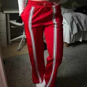Röda vida byxor med ränder - Röda byxor i träningsmaterial med vita och svarta ränder längs benen. Byxorna har resår och snöre att spänna vid midjan samt sidofickor❤️