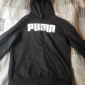 Svart hoodie från Puma - Svart hoodie från Puma med stor vit logga på bröstet. Klassisk känguruficka framtill och huva. Perfekt för en avslappnad stil.