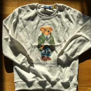 Vit sweatshirt med Polo Bear från Ralph Lauren - Vit sweatshirt från Polo Ralph Lauren med det klassiska Polo Bear-motivet framtill. Tröjan har rund halsringning och långa ärmar. Perfekt för dig som gillar streetstyle och vill ha något ikoniskt i garderoben.