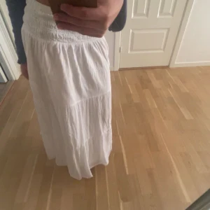 Vit långkjol från Gina Tricot - Säljer en vit långkjol från Gina Tricot med smock i midjan och lätt, flowigt tyg. Kjolen har flera volanger och är perfekt för varma dagar. Enkel att matcha med olika toppar för en fräsch look.