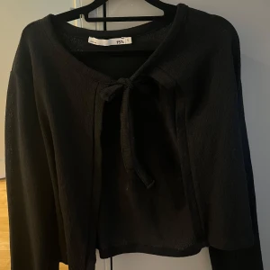 Svart knytkofta från 157 - Svart kofta från 157 i storlek S. Modellen är croppad med långa ärmar och har en knytning framtill vid halsen. Perfekt att slänga över en topp för en enkel och stilren look. Sänker i pris då den är oanvänd utan prislapp