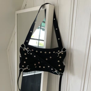 Svart axelväska med nitar och remmar oanvänd  - Cool svart axelväska med silvriga nitar och dekorativa remmar framtill. Väskan har en unik form och justerbar axelrem. Perfekt för dig som vill sticka ut med din stil.