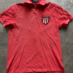 Röd piké t-shirt från Polo Ralph Lauren - Röd piké t-shirt från Polo Ralph Lauren med broderad sköld på bröstet och stor örn- och flaggdetalj på ryggen. Klassisk krage med två knappar och korta ärmar. Perfekt för en sportig och avslappnad stil. Finns lite små  slitna ställen på kragen och längs ner. 