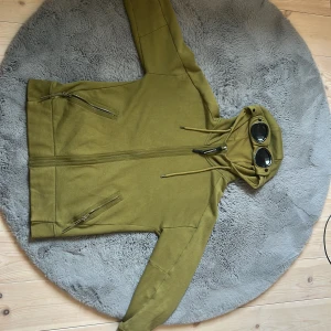 Olivgrön hoodie med glasögon från C.P. Company - Säljer en olivgrön hoodie från C.P. Company med dragkedja och ikoniska glasögon i huvan. Jackan har två fickor med dragkedja och justerbar huva med snörning. Perfekt för dig som vill sticka ut med en unik streetwear-look.