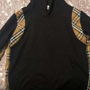 Burberry Hoodie  - Säljer en svart Burberry hoodie med klassiska beige, svarta och vita rutiga paneler längs ärmar och axlar. Tröjan har huva och magficka, samt en unik knyt-detalj på ena ärmen. 