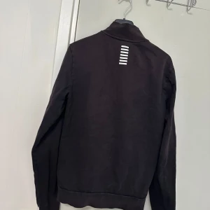 Svart zip-jacka från EA7 Emporio Armani - Svart jacka med dragkedja från EA7 Emporio Armani. Hög krage, två fickor framtill och vit logga på bröstet. Snygg detalj med vita ränder på ryggen upptill. Perfekt för en sportig och stilren look.