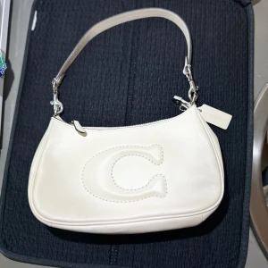 Vit axelväska från Coach - Säljer en oanvänd stilren vit axelväska från Coach med ett stort C broderat på framsidan. Väskan har silverfärgade metalldetaljer, avtagbar axelrem och dragkedja upptill. Perfekt för dig som gillar klassisk och enkel design. Långt band medföljer även. Säljer för 2000 vid snabbaffär!