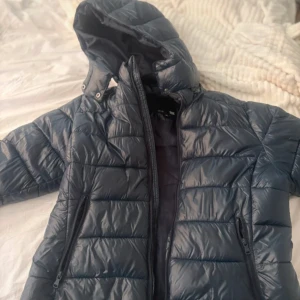 Mörkblå pufferjacka med huva - Säljer en mörkblå pufferjacka med huva och dragkedja framtill. Jackan har två sidofickor med dragkedja och quiltad design. Perfekt för kyligare dagar och enkel att matcha med olika outfits.
