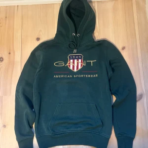 Mörkgrön hoodie från GANT - Säljer en snygg mörkgrön hoodie från GANT i bra skick. Tröjan har en broderad logga, huva med dragsko samt en stor och praktisk magficka. Stilren och bekväm – perfekt för både vardag och avslappnade tillfällen.  Hör av dig vid frågor eller om du vill ha fler bilder😁