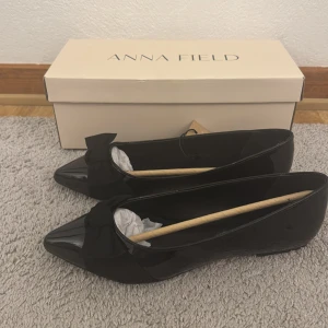 Svarta ballerinaskor med rosett från Anna Field - Snygga svarta ballerinaskor från Anna Field med lackad finish och stor rosett framtill. Skorna har spetsig tå och platt sula, perfekta för en stilren look. Kommer i originalkartong. Aldrig använd mer än att prova, jag brukar vanligtvis ha storlek 38 men anser att detta är större. 