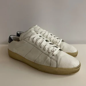 Vita sneakers från Saint Laurent - Klassiska vita sneakers från Saint Laurent med lågt skaft, vit ovandel i skinn och kontrastdetalj i svart på hälen. Skorna har snörning och en platt, beige sula. Perfekta nu för sommaren. Skriv till mig om du undrar något?