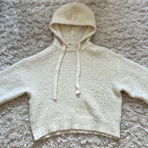 Mysig stickad hoodie i offwhite - Supermjuk och fluffig stickad hoodie i offwhite med huva och snörning. Tröjan har en croppad passform och långa ärmar, perfekt för kyliga dagar. Materialet känns riktigt cozy och ger en avslappnad vibe.