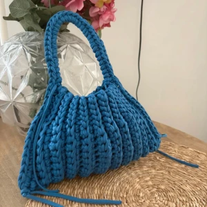 Blå stickad handväska med snörning - Superfin blå handväska i grovt stickat material med cool snörning på sidorna. Väskan har en rundad form och ett rejält handtag.