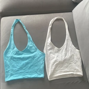 Två-pack  croppade linne-toppar i blått och vitt - Säljer två croppade linne-toppar med halterneck och djup urringning. En är ljusblå och den andra är vit. Båda är ärmlösa och i mjuk bomull, perfekta för varma dagar eller att styla lager på lager. Snygga till jeans eller kjol för en chill look.