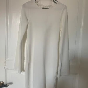 Vit långärmad klänning med draperad rygg - Stilren vit klänning med långa ärmar och snygg draperad ryggdetalj med knytband. Klänningen har en rak passform och är tillverkad i ett mjukt, lätt strukturerat material. Perfekt för dig som gillar minimalistisk och elegant stil. Köparen betalar frakt 