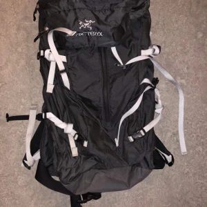 Svart ryggsäck Arc'teryx Alpha 35 Vintage Arc’teryx Hiking Backpack (OS)+ Waterproof Haglöfs - Grym svart ryggsäck från Arc'teryx, modell Alpha 35, med vita remmar och flera spännen för att justera passformen. Tillverkad i slitstark syntet och medföljer regnskydd från Haglöfs. Perfekt för äventyr och outdoor.