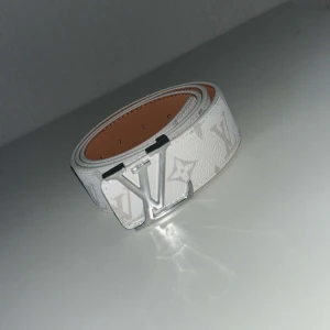 Vitt Louis Vuitton bälte i skinn - Snyggt vitt bälte från Louis Vuitton med klassiskt monogrammönster och stor LV-spänne i silver. Bältet är gjort i skinn och har en stilren, modern look som lyfter vilken outfit som helst.