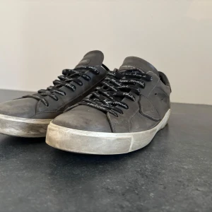 Grå Philippe Model sneakers i skinn - Snygga grå sneakers från Philippe Model i skinn med svarta snören med vit text. Klassisk låg modell med vit sula och rund tå. Perfekta för en avslappnad och trendig look.