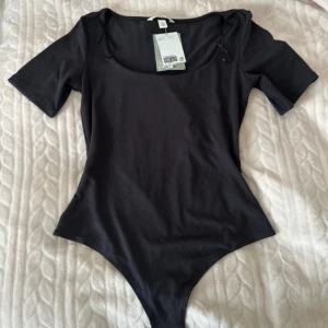 Svart snygg  bodysuit från H&M - Svart snygg  bodysuit från H&M med rundad halsringning och korta ärmar. Gjord i mjuk bomullsmix som sitter tight och smickrande. Perfekt att styla med jeans eller kjol för en clean look.