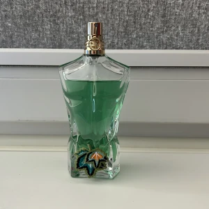 Jean Paul Gaultier Le Beau Paradise Garden - Ikonisk parfymflaska formad som en manlig torso med grön vätska och guldfärgad spraykork. Flaskan har en broderad detalj med färgglada blad vid basen. Perfekt för dig som gillar unika och snygga flaskor.