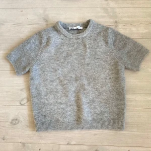 Grå stickad kortärmad tröja från Zara - Har använt några fåtal gånger. Mysig grå stickad tröja från Zara med korta ärmar och rund hals. Tröjan har en enkel och clean look med ribbade muddar vid ärmslut och nederkant. Perfekt att styla med jeans eller kjol för en chill vibe. Den ser lite konstig ut nu men är super fin på, skriv om du vill ha fler bilder.