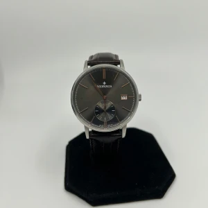 Morris herrklocka - Stilren klocka från Morris med mörkbrun läderrem och silverfärgat boett. Urtavlan är grå med diskreta index och datumvisning. Klassisk rund form och elegant design som passar till många outfits. Levereras med originalask.