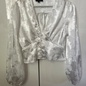 Vit blommig blus från In The Style - Superfin vit blus i satinliknande material från In The Style. Blusen har blommigt mönster, v-ringning, knappar framtill och långa puffärmar. Croppad passform som ger en trendig look. Perfekt för dig som vill sticka ut med eleganta detaljer.