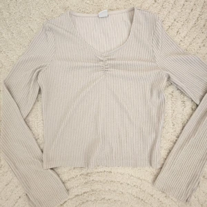 Glittrig ribbad topp från Lindex - Superfin ljusbeige långärmad topp från Lindex med glittriga silverränder. Toppen har v-ringning med rynkdetalj framtill och är ribbad i materialet. Ärmarna har en liten slits vid mudden för extra detalj. Perfekt för dig som gillar lite extra shine!