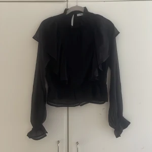 Svart volangblus  - Svart tunn (lite genomskinlig i tyget) blus från NAKD med volanger. Sömmen i midjan ger en liten peplum effekt!
