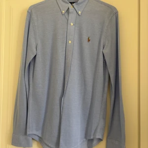 Blå oxfordskjorta från Ralph Lauren - Snygg ljusblå oxfordskjorta från Ralph Lauren med klassisk button-down krage och broderad logga på bröstet. Skjortan har långa ärmar, vita knappar och är tillverkad i bomull. Perfekt för en clean och stilren look.