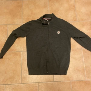 Mörkgrå kofta från Moncler med dragkedja - Snygg mörkgrå kofta från Moncler med hel dragkedja framtill och klassisk logga på bröstet. Tröjan har lång ärm, ribbade muddar och stårkrage. Perfekt för dig som gillar stilrena och exklusiva plagg i garderoben.