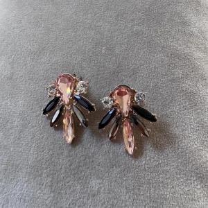 Glittriga örhängen med rosa stenar - Statementörhängen med stora droppformade rosa stenar, svarta och klara kristaller i en cool solfjäderform. Snyggt guldigt metallfäste och glittriga detaljer som verkligen drar blickarna till sig. Perfekta för att lyfta vilken outfit som helst.