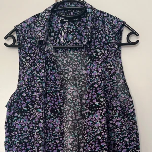 Blommig ärmlös blus från Sinsay - Säljer en luftig och ärmlös blus från Sinsay med svart botten och små blommor i lila, turkos och vitt. Blusen har krage och knäppning framtill, perfekt för dig som gillar färg och mönster. Passar till både jeans och kjol!