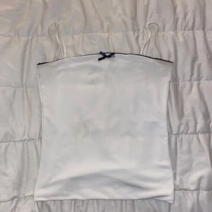 Vit bandeau topp med svart rosett BikBok - Supersöt vit bandeau topp från BikBok med tunna axelband och svart spetskant upptill. Framtill sitter en liten svart rosett som extra detalj. Toppens material känns mjukt och har en lätt glans. Perfekt för dig som gillar minimalistisk men ändå gullig stil.