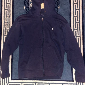 Mörkblå hoodie från Polo Ralph Lauren - Mörkblå hoodie från Polo Ralph Lauren med dragkedja framtill och klassisk vit broderad logga på bröstet. Tröjan har huva, känguruficka och är tillverkad i mjuk bomullsmix. Perfekt för en chill och stilren look.