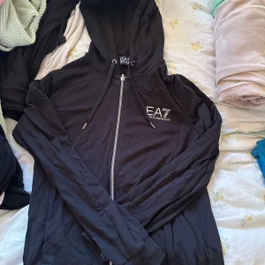 Svart hoodiejacka från EA7 Emporio Armani - Svart hoodiejacka från EA7 Emporio Armani med dragkedja framtill och snörning i huvan. Jackan har ribbade muddar och logga på bröstet. Perfekt för en sportig och avslappnad stil. (Gammalt pris 1755kr)