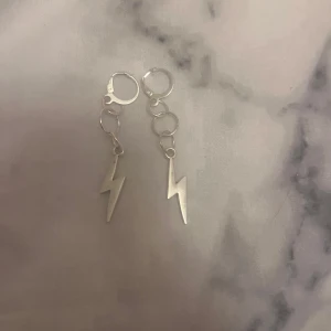 Silvriga blixtörhängen - Ett par coola örhängen i silverfärg med hängande blixtformade berlocker. De har små ringar som länkar ihop delarna och ger ett trendigt uttryck. Perfekta för dig som vill sticka ut med en edgy detalj i din stil.