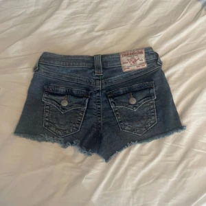 True Religion jeansshorts med knappar - Snygga blå jeansshorts från True Religion med kant och flera silverfärgade knappar framtill. Shortsen har klassiska bakfickor med fint mönster och logga på baksidan. Perfekta för sommaren och ger en bra vibe!