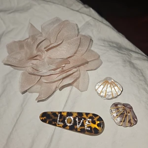 Håraccessoarer med LOVE och snäckor - Säljer ett set med en stor beige tygblomma, ett hårspänne i brun leopardmönstrad plast med texten LOVE i glittriga stenar samt två snäckformade clips i pärlemor och guld. Perfekt för att piffa upp frisyren med en cool och söt touch.