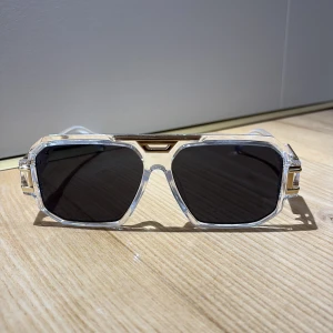 Transparenta solglasögon med guldiga detaljer - Snygga solglasögon med transparent båge och rektangulära svarta glas. Guldiga metalldetaljer vid gångjärnen och ovansidan ger en lyxig känsla. Ramen är kantig och modern, perfekt för dig som vill sticka ut med din stil. Pris 175 plus frakt.