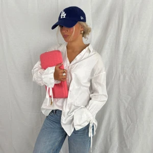 Vit oversized skjorta med snörning - Vit oversized skjorta i bomull med långa ärmar och breda manschetter med snörningsdetalj. Klassisk krage och knäppning framtill. Skjortan har en avslappnad passform och är perfekt att styla med jeans för en cool look.